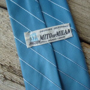 May include: A light blue tie with white diagonal stripes. The label reads "MITO of MILAN MILANO - MIAMI" and "DESIGNER SELECTIONS". The tie is made of a smooth fabric.