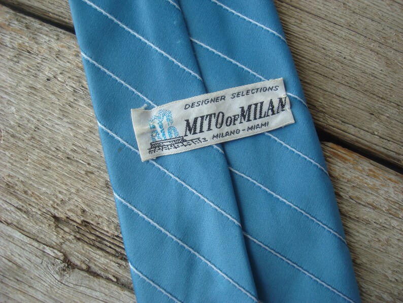 May include: A light blue tie with white diagonal stripes. The label reads "MITO of MILAN MILANO - MIAMI" and "DESIGNER SELECTIONS". The tie is made of a smooth fabric.