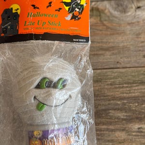 May include: A Halloween Lite Up Stick in a clear plastic package. The stick is shaped like a mummy with green eyes and a green tongue. The packaging is orange with a witch graphic and the words "Halloween Lite Up Stick".