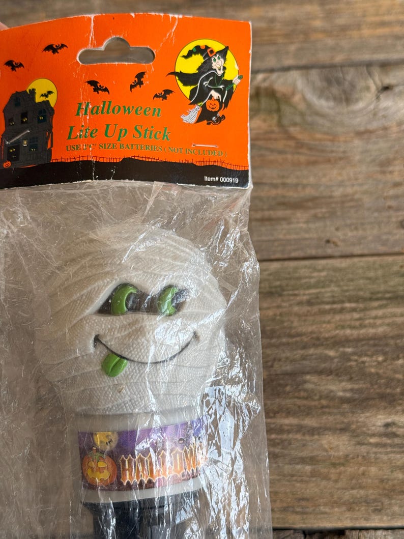 May include: A Halloween Lite Up Stick in a clear plastic package. The stick is shaped like a mummy with green eyes and a green tongue. The packaging is orange with a witch graphic and the words "Halloween Lite Up Stick".