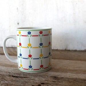 K&ouml;nnte beinhalten: Wei&szlig;e Keramiktasse mit einem sich wiederholenden Muster aus gr&uuml;nen Stielen, roten, blauen und gelben Blumen.