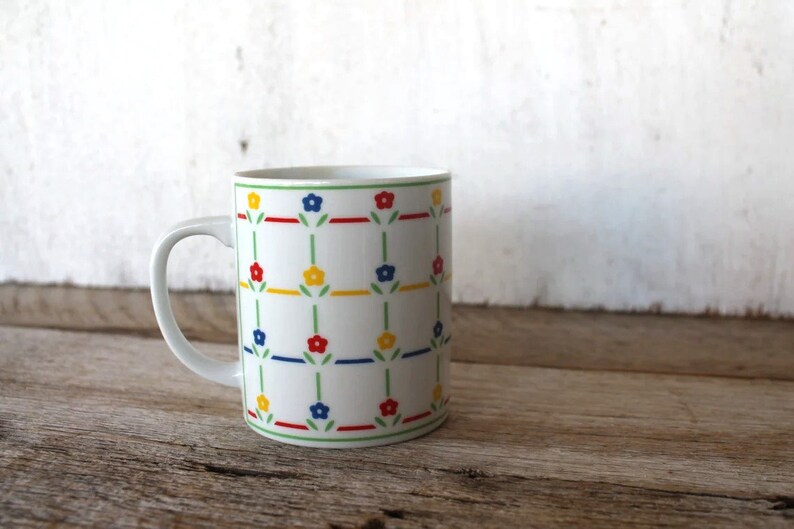 K&ouml;nnte beinhalten: Wei&szlig;e Keramiktasse mit einem sich wiederholenden Muster aus gr&uuml;nen Stielen, roten, blauen und gelben Blumen.