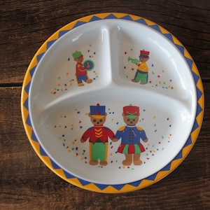 Könnte beinhalten: Ein weißer Teller mit Unterteilung und einem gelb-blauen Muster am Rand. Der Teller zeigt Illustrationen von Teddybären in Marschkapellenuniformen mit Konfetti. Der Teller ist für Kinder konzipiert.