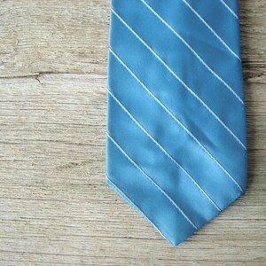 May include: A blue tie with white diagonal stripes. The tie is made of a smooth fabric and is displayed against a light wood grain background. The tie's design is simple and classic.