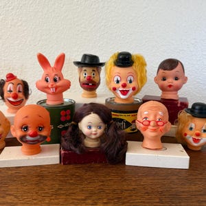 May include: A collection of vintage clown and character heads, including a rabbit, a doll, and several clowns with painted faces and wigs. The heads are displayed on a wooden surface.