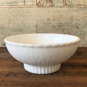 Puede incluir: Un cuenco de cerámica blanca con un patrón acanalado vertical texturizado. El cuenco tiene un borde festoneado y se asienta sobre una base de pedestal. El cuenco tiene aproximadamente 15 cm de diámetro y 10 cm de altura.