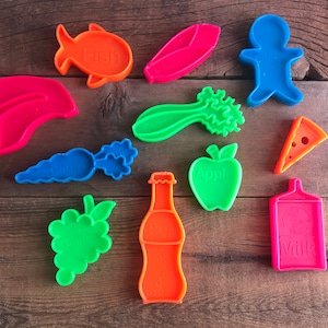 May include: A set of 10 colorful plastic cookie cutters shaped like various foods, including a fish, corn, carrot, celery, apple, grapes, soda bottle, milk carton, cookie, and a slice of cheese. Each cutter has the name of the food printed on it.