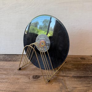May include: A gold-colored metal record holder with a black vinyl record. The record holder has a wireframe design and is sitting on a wooden surface. The record has a label that reads "SIDE 1".