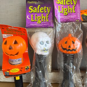 May include: A collection of Halloween-themed flashing safety lights. The lights include a pumpkin, a skull, and a pumpkin-shaped light. The packaging features the words "Flashing Safety Light" and "Halloween Pumpkin Light".