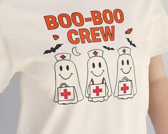May include: Cream-colored t-shirt with the text "BOO-BOO CREW" in orange. The design features three smiling ghost figures dressed as nurses, each holding a medical bag with a red cross. Bats and a crescent moon are also included.