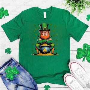 May include: Green t-shirt featuring a St. Patrick's Day design. The graphic depicts a leprechaun with a red beard and green hat, sitting in front of a pot of gold. Shamrock accents are scattered around the design. The shirt is paired with denim jeans and white sneakers.