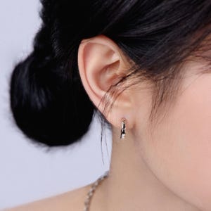 May include: Close-up of a silver hoop earring with a geometric design. The earring is worn on the ear of a person with dark hair pulled back. The earring has a small, rectangular shape with a polished finish.