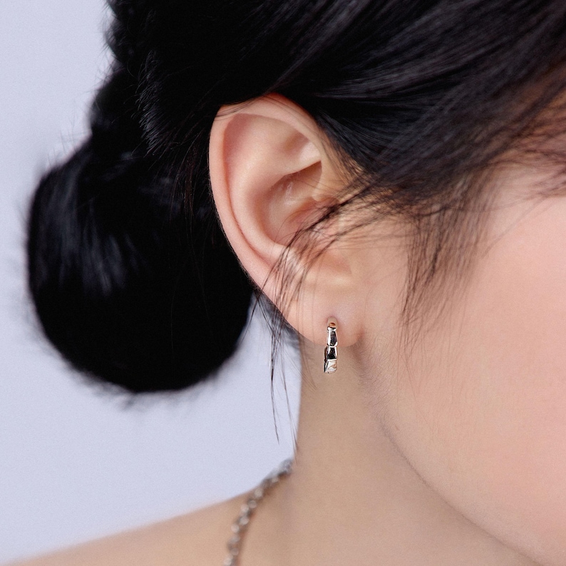 May include: Close-up of a silver hoop earring with a geometric design. The earring is worn on the ear of a person with dark hair pulled back. The earring has a small, rectangular shape with a polished finish.