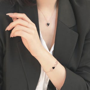 以下が含まれることがあります： 黒いハートのペンダントが付いたローズゴールドのネックレスとブレスレットのセット。ネックレスには1つのハートペンダントがあり、ブレスレットには複数のハートチャームが付いています。ジュエリーは白いシャツと黒いブレザーの上に着用されています。