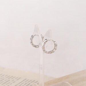 May include: A pair of silver hoop earrings with a segmented design. The earrings are displayed on clear acrylic stands against a soft, neutral background. The hoops have a modern, minimalist aesthetic.
