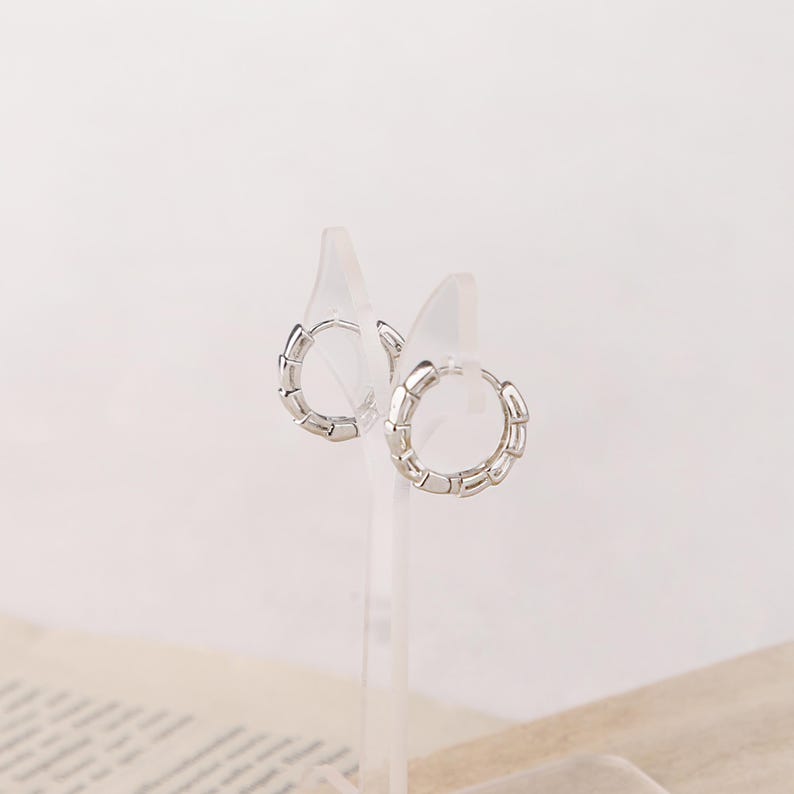 May include: A pair of silver hoop earrings with a segmented design. The earrings are displayed on clear acrylic stands against a soft, neutral background. The hoops have a modern, minimalist aesthetic.