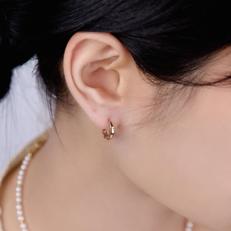 May include: A small, rose gold hoop earring is worn on the ear. The earring has a geometric design with multiple facets. The person's hair is dark, and a pearl necklace is visible.