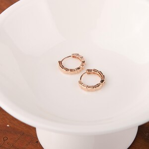 May include: Two rose gold hoop earrings with a geometric design. The earrings are small and round, with a series of rectangular segments forming the outer edge. They are displayed on a white, pedestal-style dish.