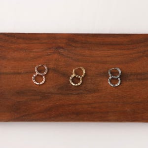 May include: Three pairs of hoop earrings in gold, silver, and rose gold tones. The earrings have a textured, chain-like design. The earrings are displayed on a dark wooden surface.