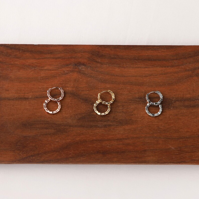 May include: Three pairs of hoop earrings in gold, silver, and rose gold tones. The earrings have a textured, chain-like design. The earrings are displayed on a dark wooden surface.