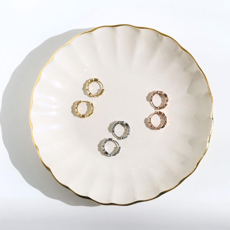 May include: A collection of hoop earrings in gold, silver, and rose gold tones, arranged on a white, scalloped-edge plate with a gold rim. The earrings have a segmented design, and the lighting creates soft shadows.