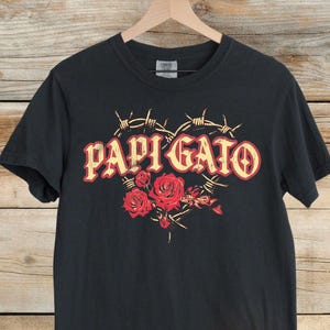 May include: Black t-shirt with the text "PAPI GATO" in red and yellow lettering, surrounded by barbed wire and red roses. The shirt is hanging on a wooden hanger against a wooden background.