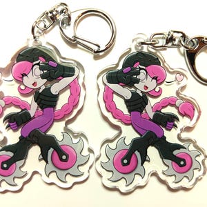 May include: Two pink and black character keyrings with a pink heart and a silver chain. The character is wearing a black helmet, black gloves, and purple trousers. The character is riding on a pair of pink wheels with black blades.