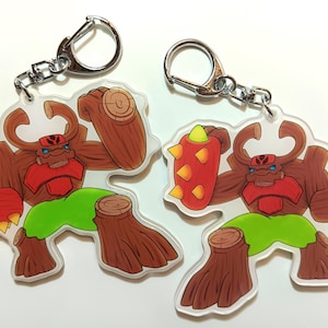 May include: Two brown and green cartoon character keychains with a red and yellow design. One keychain has a spiked ball and the other has a log.
