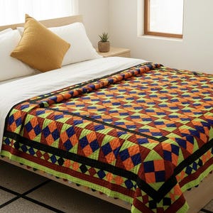 Op de afbeelding: Een bed met witte lakens, kussens en een kleurrijke quilt. De quilt heeft een geometrisch patroon van oranje, blauwe en groene ruiten. Een mosterdkleurig kussen ligt tegen het hoofdeinde. De kamer heeft een neutraal kleurenpalet.