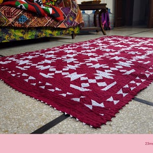 Op de afbeelding: Een rechthoekig, handgemaakt rood tapijt met een wit geometrisch patroon. Het tapijt heeft een getextureerd oppervlak en een decoratieve, zaagtandrand. Het tapijt ligt op een gevlekte tegelvloer, met een patroonbank op de achtergrond.