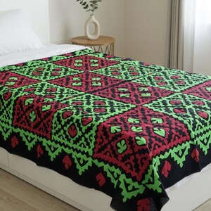 May include: A bedspread with a geometric pattern in black, red, and green. The design features diamond shapes with leaf-like details. The bedspread is on a white bed with a white pillow.