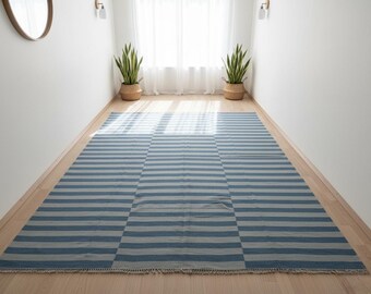 Op de afbeelding: Een rechthoekig tapijt met blauwe en grijze horizontale strepen. Het tapijt is gemaakt van geweven stof en heeft een gefranjerde rand. Het tapijt ligt op een lichtgekleurde houten vloer.