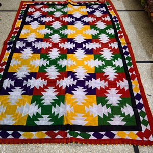 May include: A colorful, rectangular quilt with a geometric pattern. The quilt features squares of white, yellow, red, green, and purple, bordered by black and red trim. The quilt is likely handmade and suitable for a bed or wall hanging.