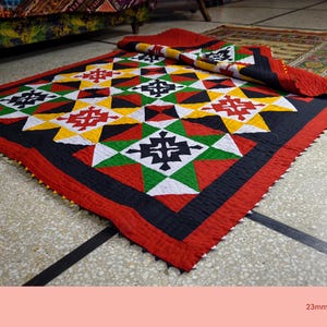 Pode incluir: Um edredom colorido com um padrão geométrico e uma borda vermelha. O edredom apresenta um design repetitivo de formas pretas, brancas, amarelas, verdes e vermelhas. O edredom está dobrado sobre si mesmo.