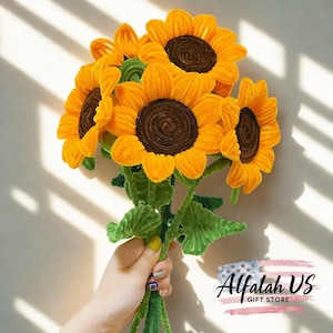 Op de afbeelding: Een boeket handgemaakte zonnebloemen met heldergele bloemblaadjes en donkerbruine centra. De stelen zijn groen en aan elkaar gebonden, met groene bladeren. Het boeket wordt in een hand gehouden. De tekst "Alfalah US GIFT STORE" staat rechtsonder.