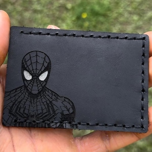 May include: Black leather wallet with a Spiderman design. The rectangular wallet has stitched edges and a Spiderman graphic etched into the leather. The Spiderman graphic is on the front of the wallet.