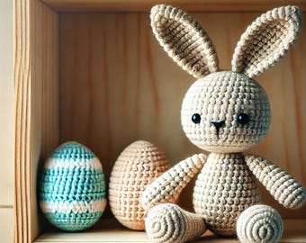 May include: A crocheted bunny rabbit toy sits on a wooden shelf with two crocheted Easter eggs. The bunny is beige and has black eyes. The eggs are blue and white and tan.