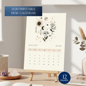 Può includere: Un calendario da scrivania stampabile del 2026 su un piccolo cavalletto di legno. Il calendario presenta uno sfondo beige con illustrazioni a tema celeste e il testo "2026 PRINTABLE DESK CALENDAR". Il calendario è esposto su una scrivania con altri oggetti decorativi.