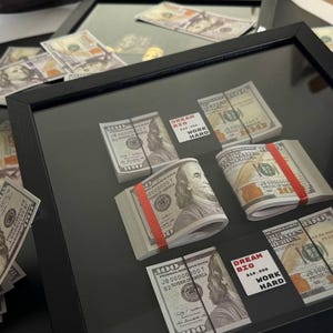 Op de afbeelding: Een ingelijst kunstwerk met stapels gesimuleerde $ 100-biljetten, elk beveiligd met een rood elastiekje. De stapels zijn in een rasterpatroon gerangschikt, met een motiverende boodschap bovenaan en onderaan: "Dream Big, Work Hard".