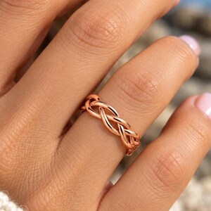 May include: A rose gold ring featuring a continuous braided or woven metal design. The band has a polished, reflective surface and a uniform width, showcasing an intricate pattern of interlocking strands that wrap around the entire circumference.