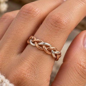 May include: A ring featuring a braided design composed of alternating strands of rose gold and silver-toned metal. The band has a continuous woven pattern that wraps around the finger, showcasing a polished finish on both the copper-hued and silver-coloured metal segments.