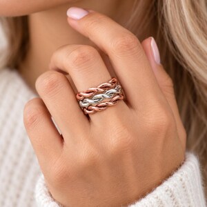 May include: A multi-band ring featuring three intertwined twisted metal strands. The design consists of two outer bands in a warm copper tone and a single central band in a bright silver tone, creating a layered and textured appearance.
