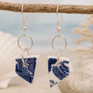 Può includere: Un paio di orecchini pendenti con frammenti triangolari di ceramica bianca a motivi blu. Ogni orecchino è sospeso a un gancio in metallo argentato e a un anello, mostrando intricati disegni floreali e paesaggistici di colore blu.