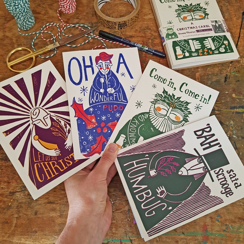 May include: Four Christmas cards with colorful illustrations and text. The cards feature a red and blue illustration of a man singing "Oh, a Wonderful Pudd", a green illustration of a man with a holly wreath saying "Come in, Come in!" a purple and green illustration of a man with a top hat saying "Bah Humbug!" and a purple and yellow illustration of a man with a sunburst behind him saying "Let us see Christ".