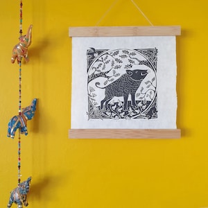 A square black & white linocut print hanging against a yellow wall alongside a string of colourful elephants. The print shows a wild boar in a circle framed tree branches, plants and other woodland animals.