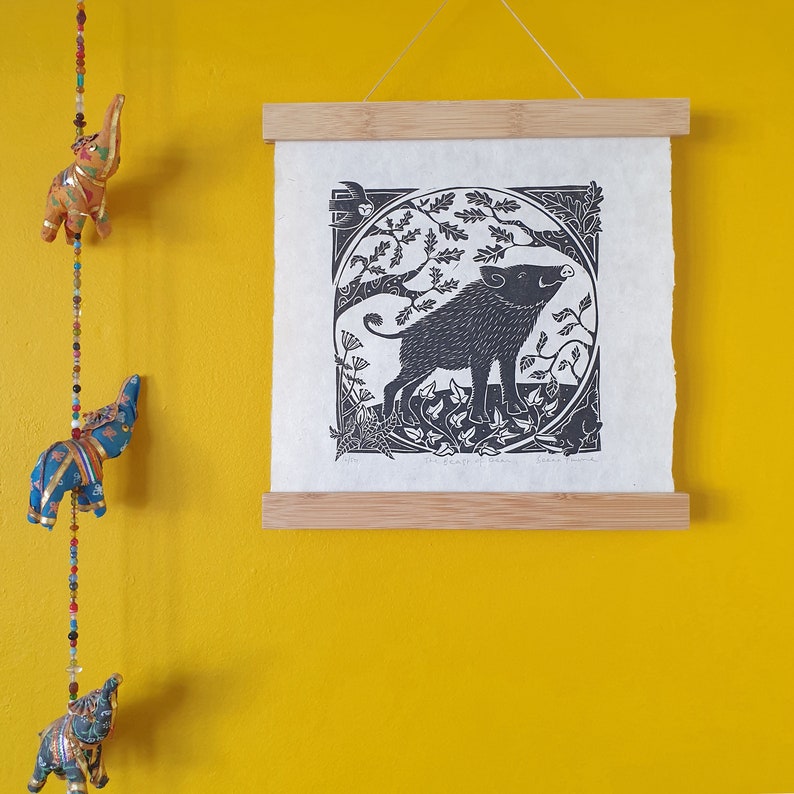 A square black & white linocut print hanging against a yellow wall alongside a string of colourful elephants. The print shows a wild boar in a circle framed tree branches, plants and other woodland animals.