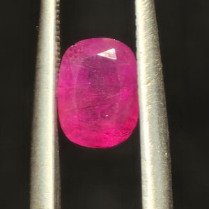 Puede incluir: Una gema de color rosa intenso con corte ovalado, sujeta firmemente entre las dos puntas metálicas de unas pinzas. La piedra presenta una superficie facetada que refleja la luz, mostrando un tono magenta profundo y saturado.