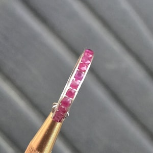 May include: A silver ring with a row of small, vibrant red gemstones. The ring is held by a gold-colored tool, showcasing the gemstones' color and the ring's design. The gemstones are evenly spaced, creating a continuous line of color.