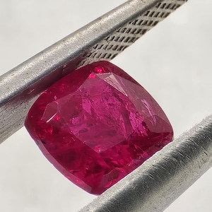 Peut inclure: Un rubis vibrant, taillé en coussin, maintenu par une pince argentée. Le rubis affiche une couleur rouge profond et saturé avec de multiples facettes, reflétant la lumière. La pince a une surface texturée.