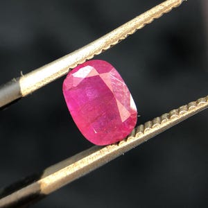 May include: A faceted, oval-shaped ruby gemstone, held by tweezers against a dark background. The ruby displays a vibrant, deep pink-red color, with multiple facets catching the light. The tweezers are a light gold color.